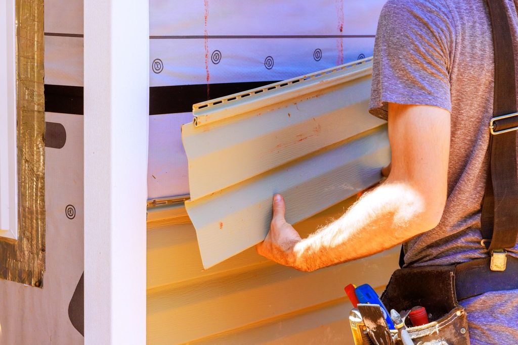 Worker holds vinyl siding panel against house wall with weather barrier visible illustrating cold-weather cladding installation